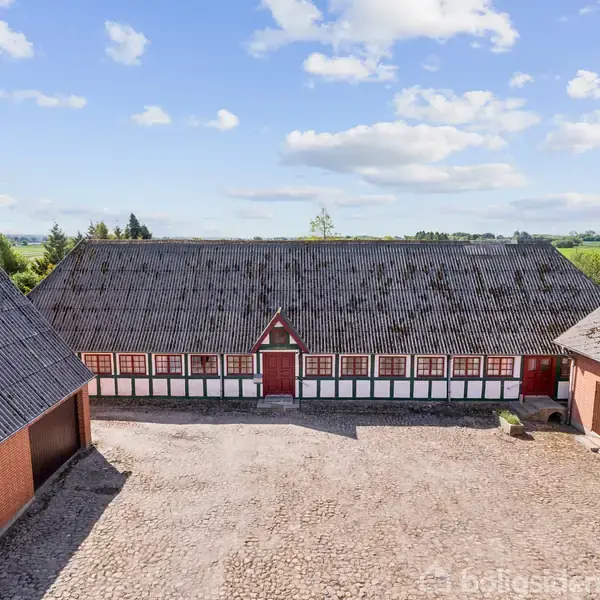 Gårdsbygning med sort tag og hvid bindingsværk, står stille mellem røde teglstensbygninger. Grusbelagt gårdsplads omgiver strukturen, mens marker strækker sig i baggrunden under en blå, let skyet himmel.