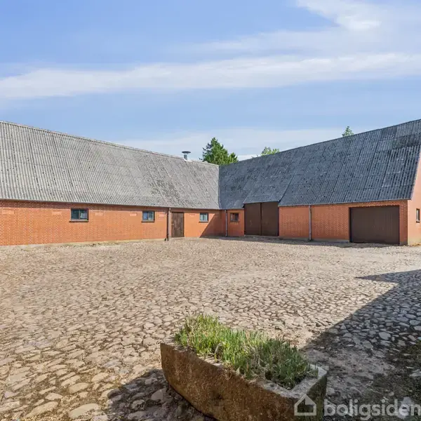 En firkantet gårdsplads med brosten omgivet af røde murstensbygninger med skrå tage. Grønt område ses i baggrunden under en blå himmel.