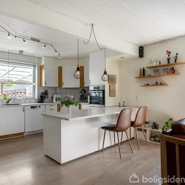 Et moderne køkken med en central hvid ø, to hængende lamper og brune stole. I baggrunden er der hvide skabe, madlavningsudstyr, og åben hylde opbevaring ved siden af en stor sofa.