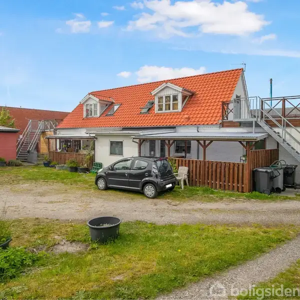 Et lille sort bil holder parkeret foran en hvid bygning med rødt tegltag. Til venstre ses en garage, og der er en åben plads med græs og grus.