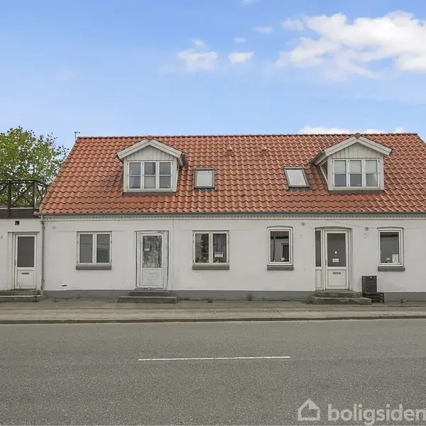 En hvid toetagers bygning med rødt tegltag står på en stille vej. Bygningen har små vinduer og døre foran, med en terrasse til venstre omgivet af grønt løv.