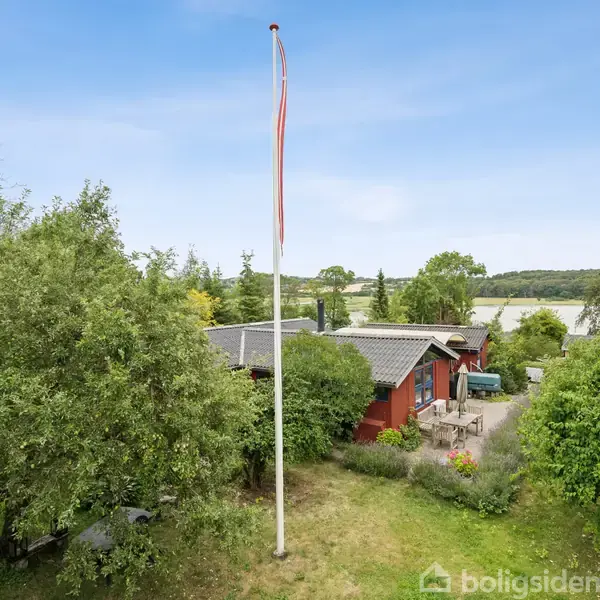 Flagstang står stille i en grøn have, omgivet af træer og buske, med et hus i baggrunden nær en sø under en klar blå himmel.