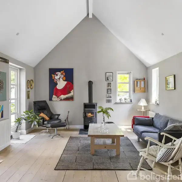 Stue med sofa, lænestol og brændeovn i centrum, omgivet af kunst på væggene. Lyse vægge, træloft og et stort vindue til venstre skaber en lys atmosfære.