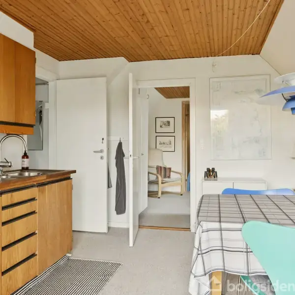 Køkken med brune skabe og sort bordplade. Spisebord med gråternet dug og turkis stole. Trævæg loft. Dør fører til en stue med lænestol og billeder på væggen. Ingen tekst.