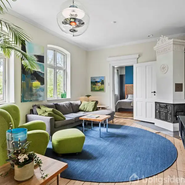 Grøn lænestol og skammel står ved et stort vindue i en stue, omgivet af sofa, bord, pejs og blå tæppe. En plante og malerier pynter rummet. Fjernsyn hænger på væggen.