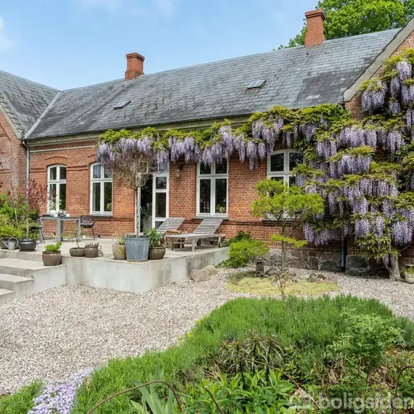 En murstensbygning med klatrende blomster pryder facaden, mens havemøbler står på en terrasse. Omgivet af en grussti og grønne planter i en have.