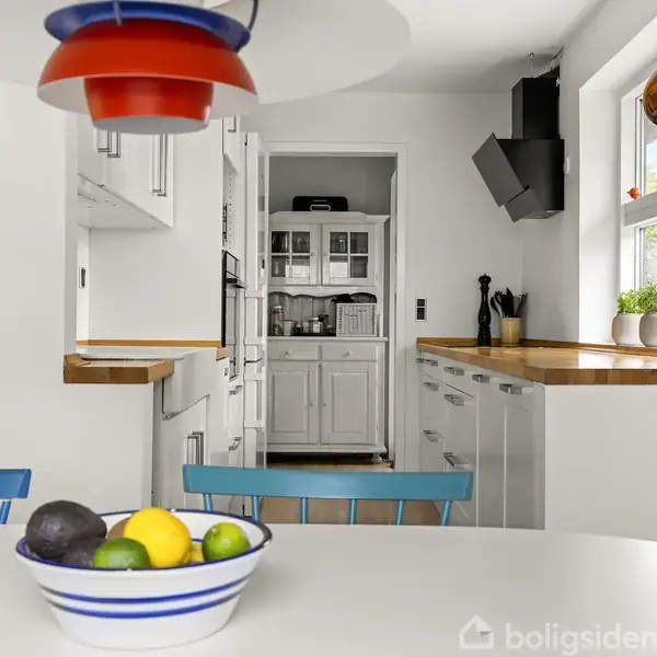 Farverig skål med frugt på hvidt bord; omkringliggende køkken med lyse skabe, træbordplader, moderne lamper og et vindue med blomster. Pæn, organiseret og lys indretning.