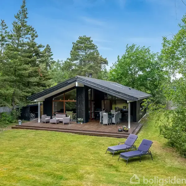 Sommerhus med sort træfacade og terrasse, indeholder udendørs siddepladser. To liggestole står på græsplænen. Omgivet af frodig have med træer.