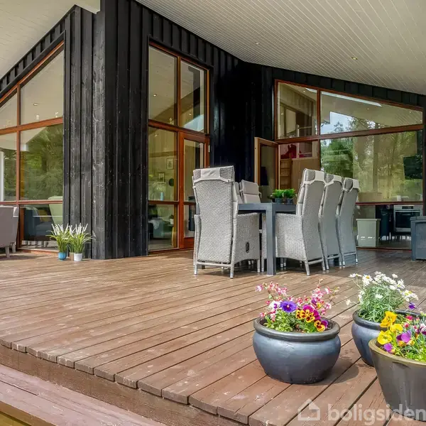 Terrasse med træmøbler og plantekrukker fyldt med farverige blomster, placeret foran et moderne træhus med store vinduer. Omgivet af natur, der skaber en fredelig atmosfære.