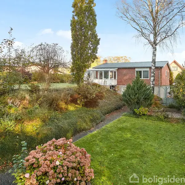 Et rødstenshus står stille omgivet af en frodig have med græsplæne, blomster og træer. I baggrunden ses flere huse og en blå himmel.
