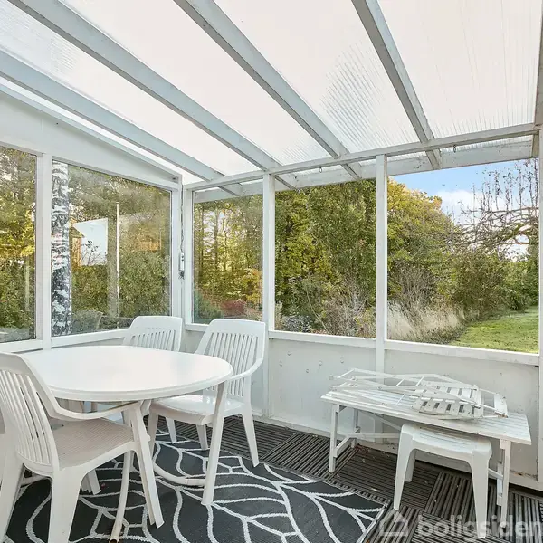 Et hvidt bord med stole står i en lys udestue, omgivet af store vinduer, der giver udsigt til en frodig have med græs og træer udenfor.