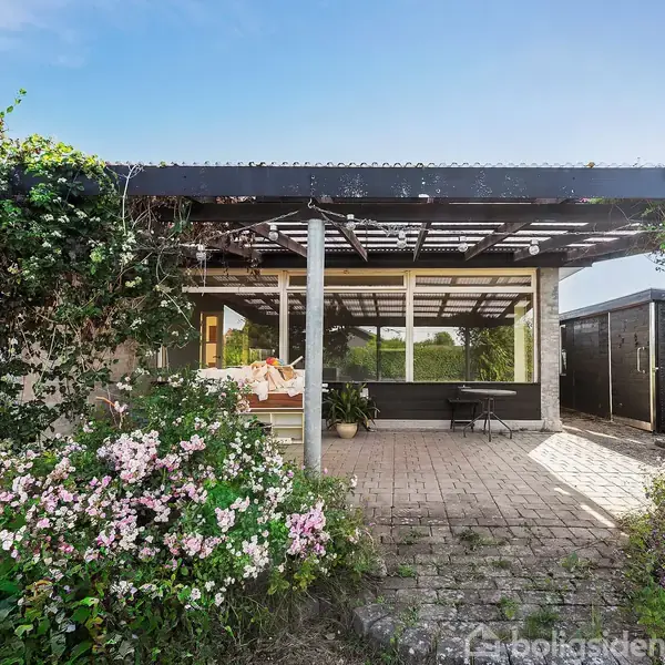 Veranda med trætage dækket af slyngplanter. Foran står et bord og en bænk på flisebelagt terrasse. Omkring er der blomstrende buske og grønne planter under en klar blå himmel.