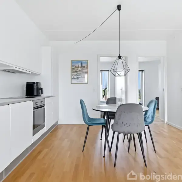 Et moderne køkken med hvide skabe og en indbygget ovn. Et rundt spisebord med blå stole står i rummet, båret af en geometrisk lampe. Trægulve og hvidmalede vægge.