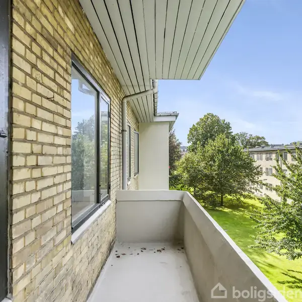 Balkon med lyse murstensvægge åbner mod en grøn have med træer og græsplæne. Bygningens facade og blå himmel ses i baggrunden.