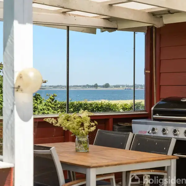 Grill står stille på en terrasse ved siden af et træbord med blomster, under et lyst tag. I baggrunden ses havudsigt og kystlandskab gennem glasvinduer.