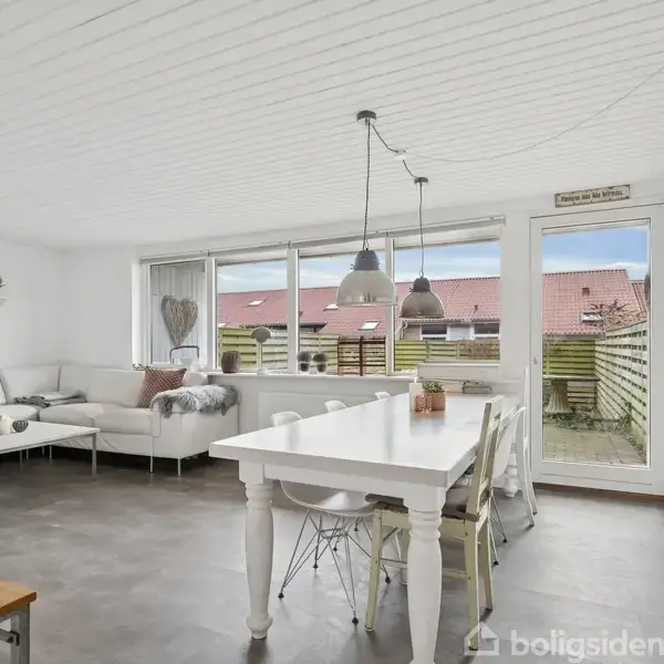 Stue med hvid sofa og stort hvidt spisebord, placeret ved siden af en glasdør, der fører ud til en terrasse. Væggene er hvide med minimalistisk indretning.