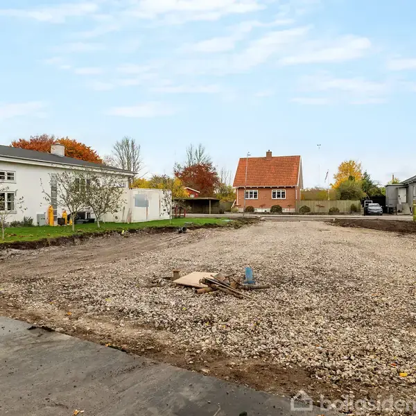 En grusbelagt byggegrund ligger i et villakvarter med huse omkring. Til venstre ses et hvidt hus og til højre et rødt hus. Det er efterår med farverige træer.
