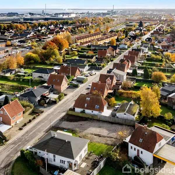 Række af huse med røde tage og haver, set fra luften. Omgivet af efterårsfarvede træer og en industriområde i baggrunden. Train tracks ses også i det fjerne.