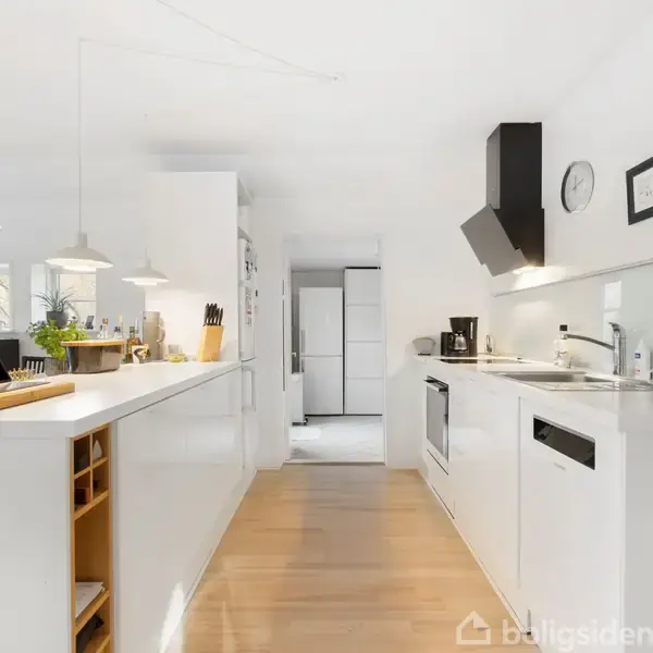 Et moderne køkken med hvide skabe og træbordplade, hvor en bærbar computer og køkkenredskaber står. Omgivelserne er lyse med hængende lamper. En væg har et ur og en indrammet plakat.