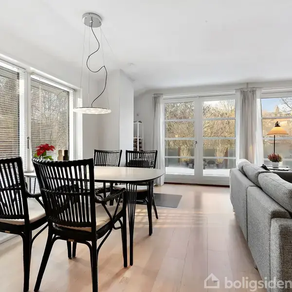 Et spisebord med sorte stole står i en lys stue med store vinduer. En grå sofa vender mod vinduerne, og en lampe står ved siden af en stol.