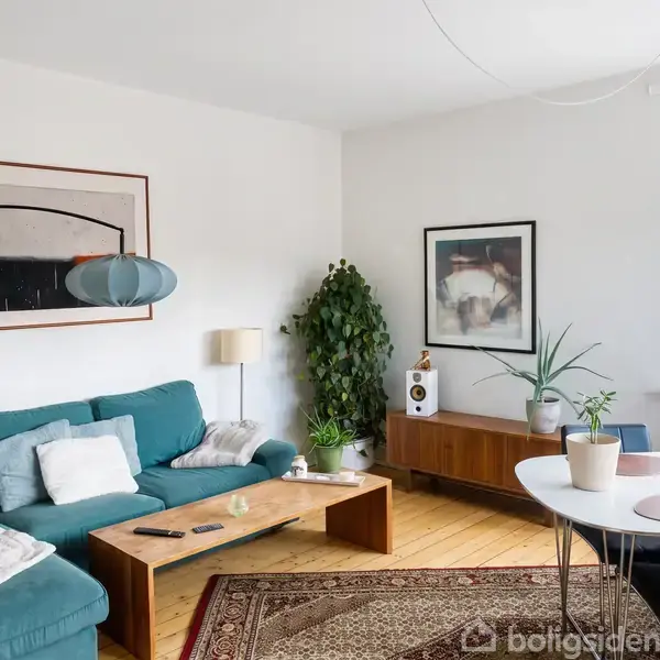 En stue med en blå sofa fyldt med puder, placeret ved siden af et lavt træbord. En stor plante står i hjørnet, mens et maleri hænger på en hvid væg.