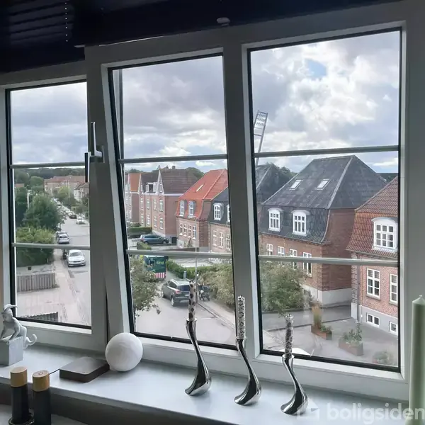 Et stort vindue åbner ud mod en gade med rækkehuse og biler. Indersiden af vinduet er dekoreret med moderne pyntegenstande og en kurv.