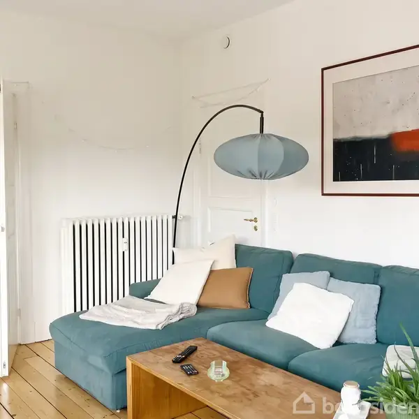 En blå sofa står i en stue med træplankegulve. Der ligger puder og tæpper på sofaen. Et sofabord med fjernbetjeninger står foran, og en stor plante ses i baggrunden.