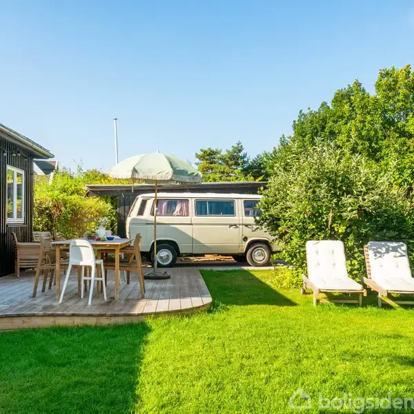 Campingvogn parkeret i have, omgivet af grønne buske og træer. Foran står et bord med stole på en træterrasse ved siden af et sort hus. Solskin over græsplænen.