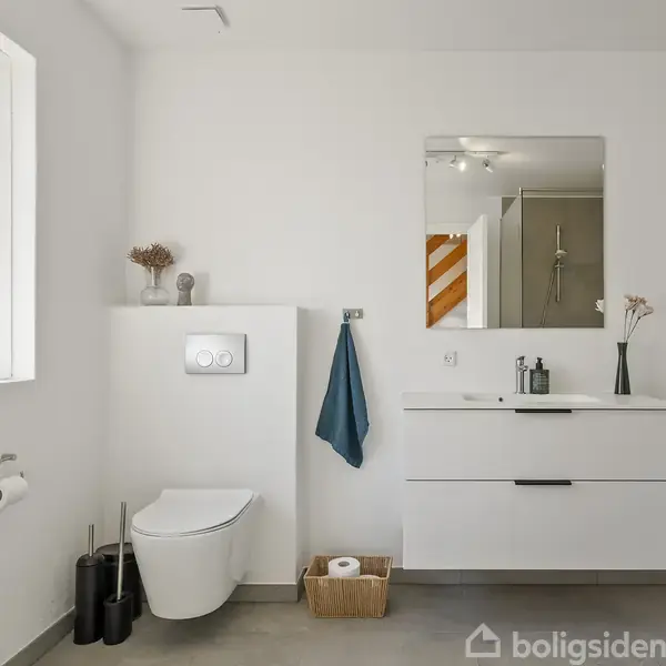 Toilet står ved siden af håndvaskskab med spejl, omgivet af hvid væg. Et blåt håndklæde hænger ved siden af, og der er en kurv og stol med billede på gulvet.