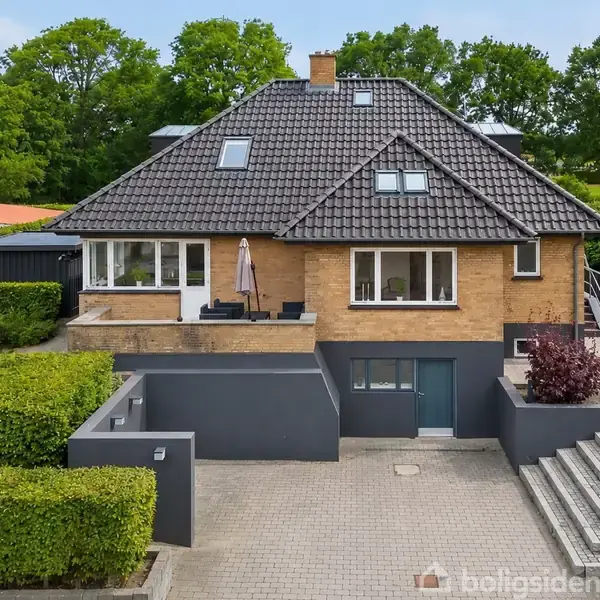 Hus med brun murstensfacade og sort tag, terrasse på første sal med parasol. Foran er der flisebelagt gårdhave og trappe, omgivet af hæk og træer i baggrunden.