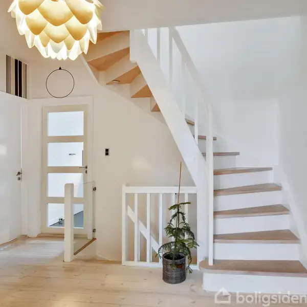 Trappeopgang i et lyst indrettet rum med hvide vægge og lyse trægulve, dekoreret med en moderne lampe og grøn plante ved gelænderet. Striped tapet til højre for døren.