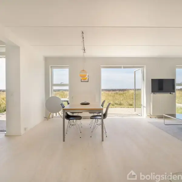 Spisebord med stole står centralt i et lyst rum. Store vinduer og døre åbner mod naturudsigt. Sofa og tv findes til højre, skaber en moderne atmosfære.