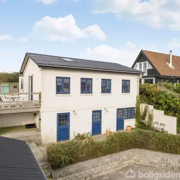 Hvid to-etagers hus med blå døre og vinduer. Huset har en træterrasse til venstre. Det ligger i et grønt område med et andet hus i baggrunden under en blå himmel.