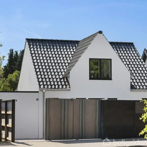 Et hvidt, moderne hus med sort tegltag står i en grøn have. Vinduerne er store og firkantede. Foran huset er der en indkørsel med en sort trælåge.