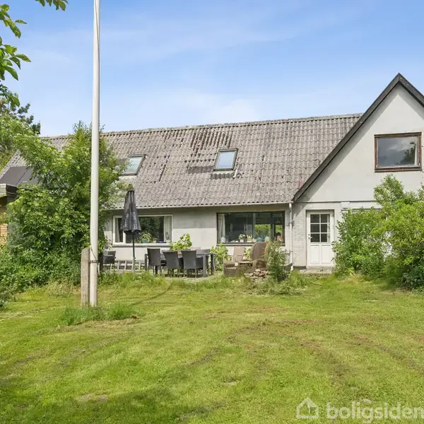 Hus med hvide vægge og gråt tag står i en grøn have med græsplæne og buske. Et flagstang står til venstre, og havemøbler ses foran huset.