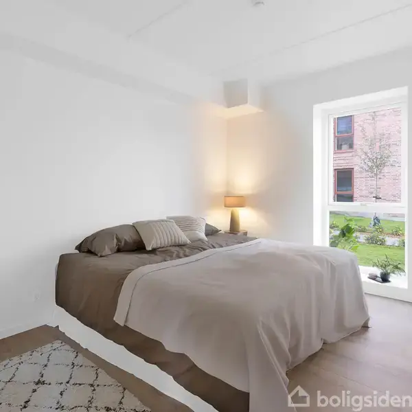 En seng med gråt sengetøj står mod en hvid væg. En tændt lampe står ved hovedgærdet. Ud af vinduet ses en græsplæne og en murstensbygning.