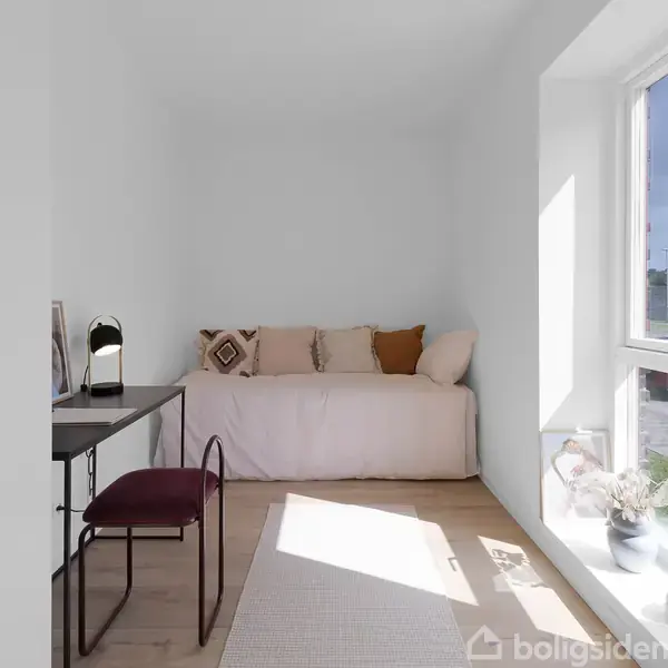 Et lille rum med en sofa fyldt med puder i hjørnet. Et skrivebord med stol står ved siden af, mens lyset skinner ind gennem et stort vindue.
