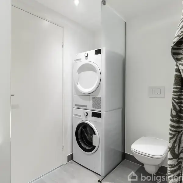 Vaskemaskine og tørretumbler er stablet, stående ved hvide vægge. Ved siden af er et væghængt toilet. Et håndklæde hænger i højre side af billedet.