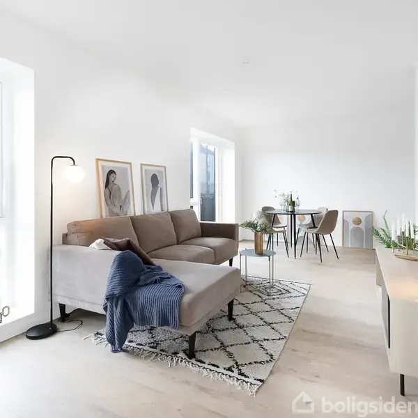 Sofa i brun farve står i en moderne stue med lyst trægulv, tæppe og vægkunst. Spisebord med stole ses nær vindue. Grønne planter pynter rummet.