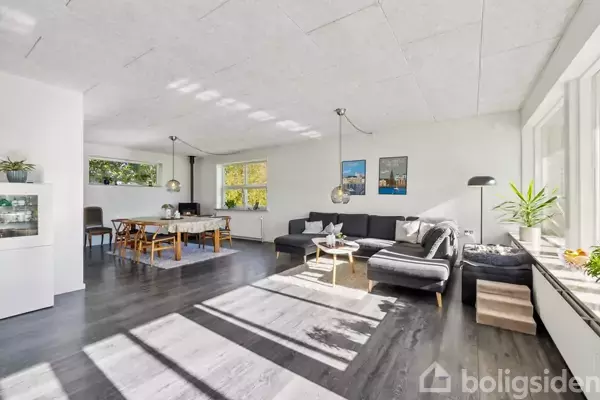 En moderne stue med mørkt trægulv og en stor, grå sofa placeret ved store vinduer, der lader naturligt lys ind. Spisebord med stole ses i baggrunden ved siden af planter.