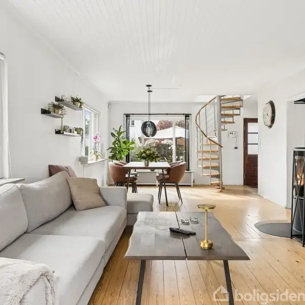 Stue med en lysegrå sofa, sofabord og væghængt TV. Spiseområde med bord og stole, tæt på spiraltrappe. Store vinduer lader naturligt lys ind, med planter som dekoration.