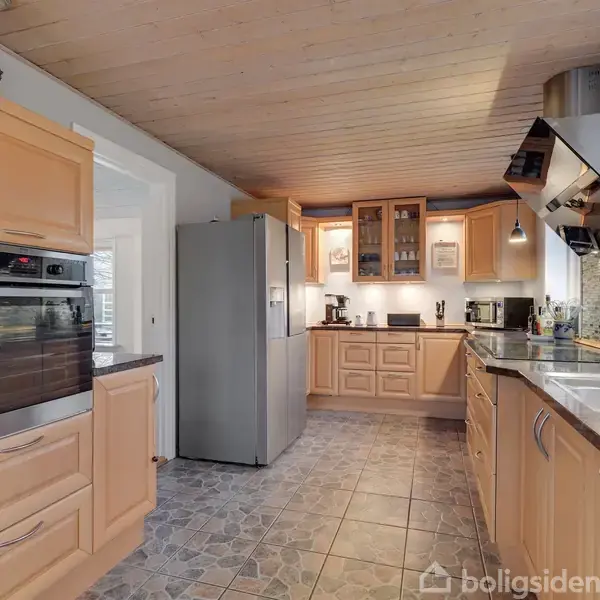 Køkken med træskabe og stålapparater. Køleskabet og indbygningsovnen står til venstre. En vask med armatur er i højre side, mens andre køkkenmaskiner står på bordet. Klinkegulv og træloft.