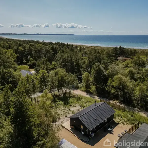 Et sort træhus står stille blandt træer i et skovområde tæt på en kystlinje med en bred strand og hav i baggrunden under en himmel med få skyer.