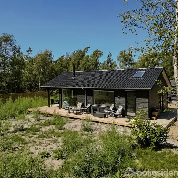 Et sort træhus står stille på en terrasse med havemøbler omgivet af græs og træer i en frodig skovområde.