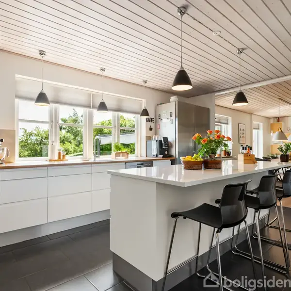 Et moderne køkken med en stor, hvid køkkenø omgivet af sorte barstole. Rummet er lyst og rummeligt med hængende lamper og grønne planter. Vinduerne viser udsigt til en have.