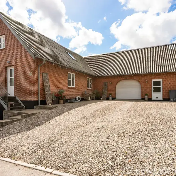 Rødstenshus med skråt tag står stille. Grusbelagt gårdsplads med trappe til hoveddøren og tilknyttet garage. Skyfri blå himmel og grønne træer danner baggrunden.