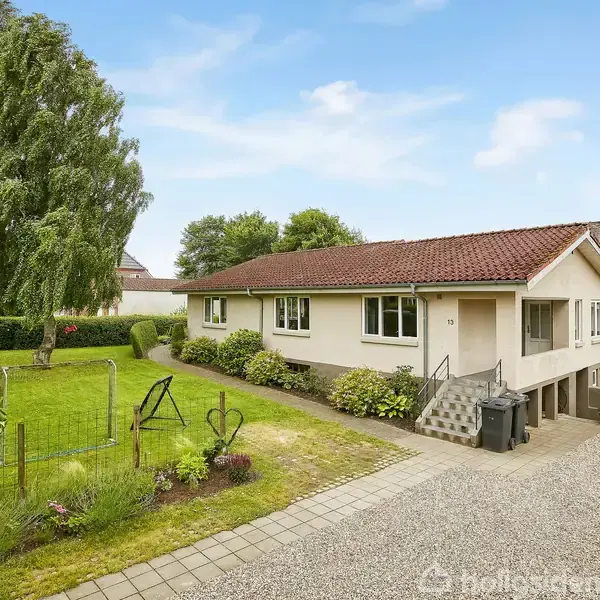 Hus med rødt tag står på en stor grund med græslagt have. Grusindkørsel fører til hoveddør og garage. Store træer og grønne buske omkranser haven.