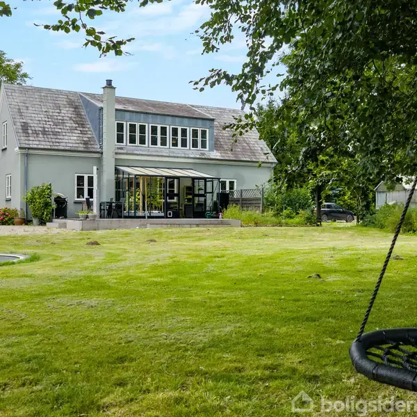 Et gråt hus med stor glasveranda står på en velholdt græsplæne. En træsving hænger fra et træ til højre, og en trampolin er til venstre på plænen.