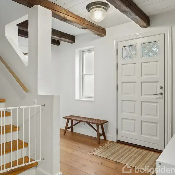 Trappe med trælister leder opad, ved siden af hvid væg, i entre med trædør. Bænk og måtte på trægulv, lysekrone i loftet og reol med tæppe til højre.