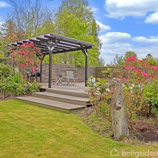 En træterrasse med et pergola-tag, der dækker et sæt havemøbler, står i en have omgivet af blomstrende buske og grønne træer under en blå himmel med lette skyer.